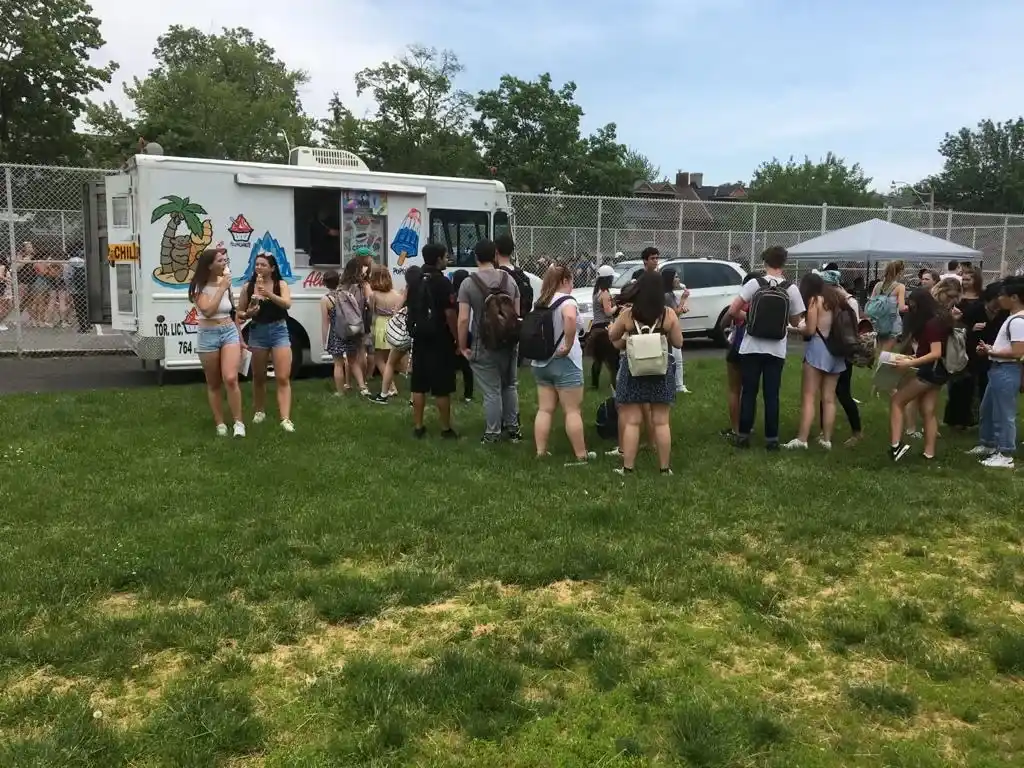 Students gathered near an ice cream truck at a school event on a grassy field, enjoying ice cream on a sunny day.