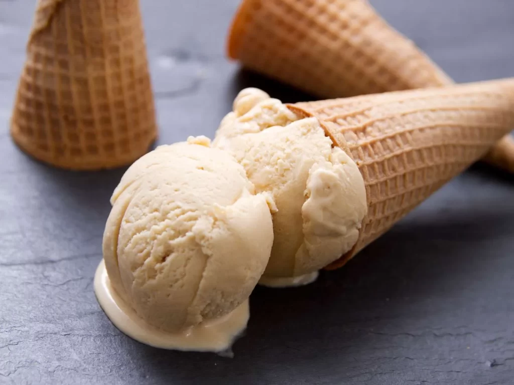 A close-up of a golden waffle cone filled with creamy vanilla ice cream, placed on a dark surface.