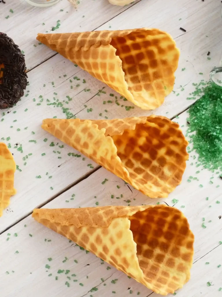 Golden waffle ice cream cones placed on a white wooden surface, surrounded by decorative sprinkles.